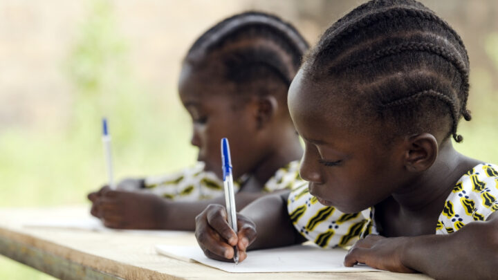African Children at School Doing Homework. African ethnicity students writing their essay in an African school. They're holding blue pens to write down their homework whilst sitting in their desk.